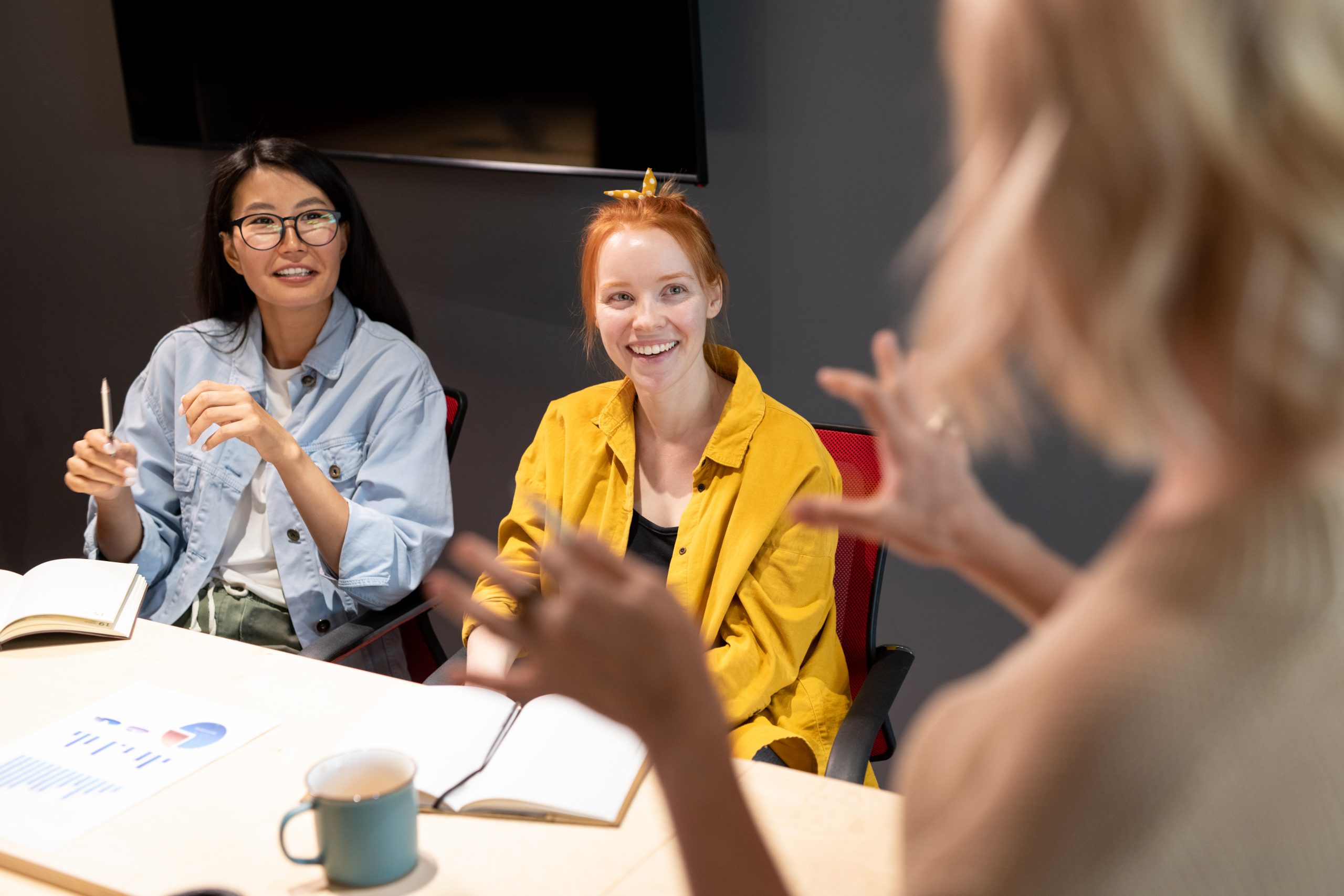 Diverse team of professionals in engaged meeting discussion, with one person raising hand to speak while others listen attentively, demonstrating active participation and presence
