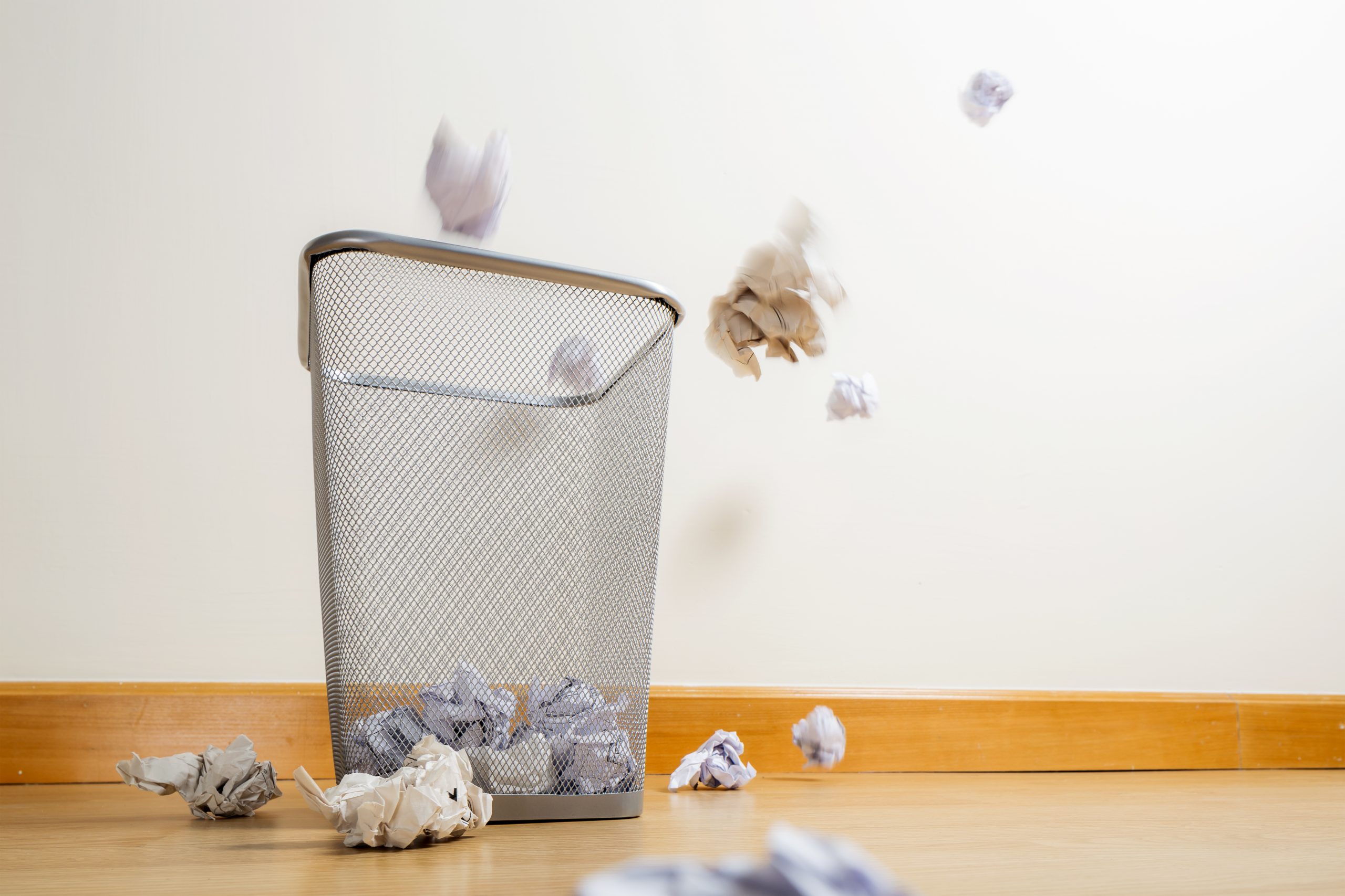 A metal mesh wastebasket filled with crumpled paper balls, with additional discarded paper scattered on the wooden floor around it, symbolizing repeated attempts and "failed" improvement initiatives