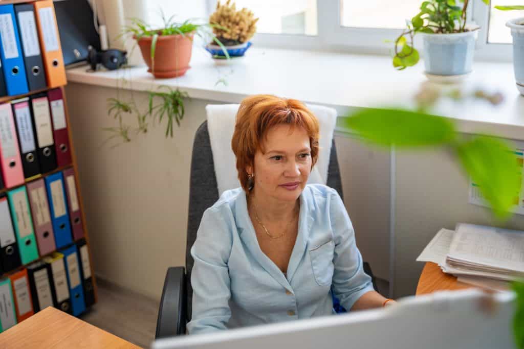 Woman at office desk looking at computer screen, surrounded by colorful binders and plants, conveying a busy, hands-on work environment