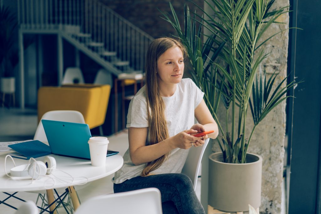 A woman sitting at a small table beside a potted plant, smiling as she looks at her phone
